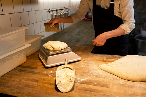 food cooking, baking and people concept - baker with bench cutter portioning and weighing bread dough on scale at bakery kitchen