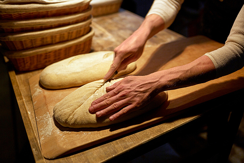 food cooking, baking and people concept - baker hands making bread and cutting dough with knife at bakery
