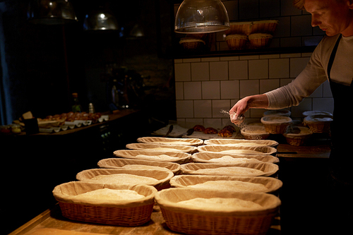 food cooking, baking and people concept - baker making bread and pouring flour to baskets while dough rising at bakery kitchen
