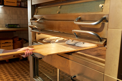 food, cooking and baking concept - baker hand with peel putting yeast dough into bread oven at bakery kitchen