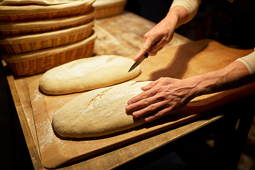 food cooking, baking and people concept - baker hands making bread and cutting dough with knife at bakery
