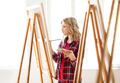 art school, creativity and people concept - student girl or artist with easel, palette and paint brush painting at studio