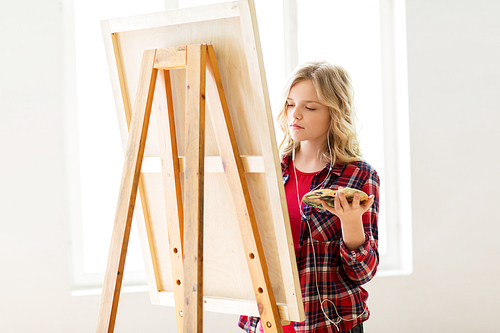 art school, creativity and people concept - student girl or artist with earphones, easel and palette painting at studio