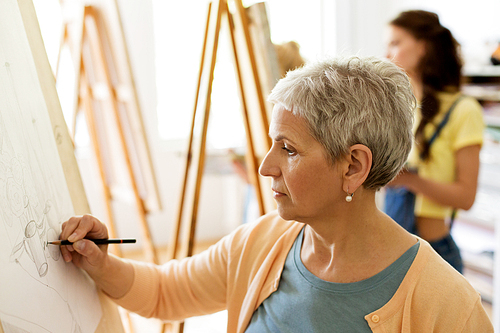 creativity, education and people concept - senior woman drawing on easel at art school studio