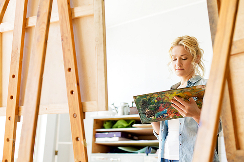art school, creativity and people concept - woman with easel and palette painting at studio