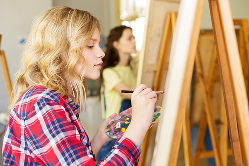 art school, creativity and people concept - teenage girl with easel, palette and brush painting at studio