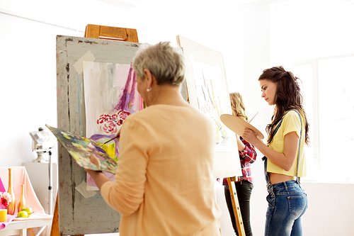 creativity, education and people concept - senior woman with brush and palette painting still life picture on easel at art school studio