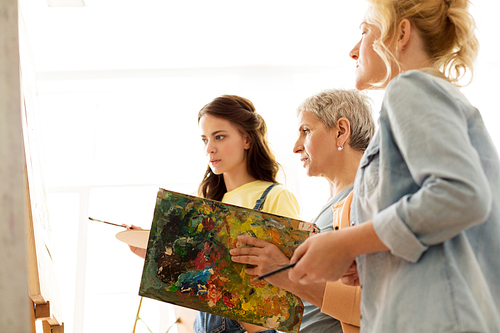 creativity, education and people concept - group of female artists or students with brushes and palettes and teacher painting on easel at art school studio
