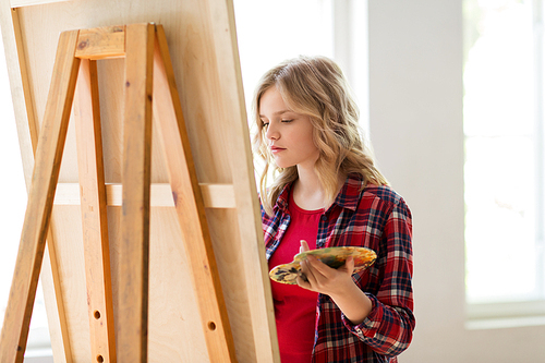 art school, creativity and people concept - student girl or artist with easel and palette painting at studio