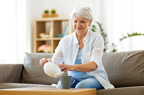 age, drink and people concept - senior woman pouring tea from teapot to cup and drinking it at home