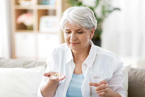 age, medicine, healthcare and people concept - senior woman with glass of water taking pills at home