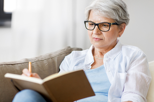 age, leisure and people concept - close up of senior woman in glasses writing to notebook or diary at home