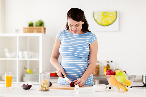 healthy eating, cooking, pregnancy and people concept - pregnant woman with knife and blender cup chopping fruits at home kitchen