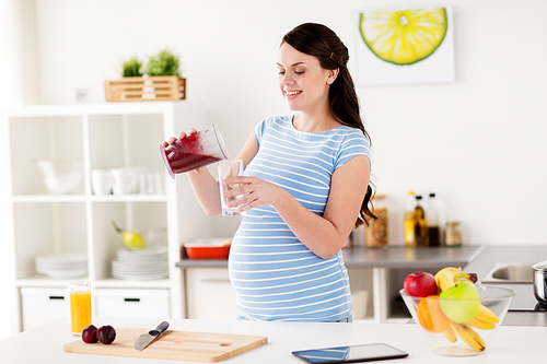 healthy eating, cooking, pregnancy and people concept - pregnant woman with blender cup pouring fruit smoothie drink into glass at home kitchen
