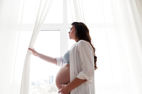 pregnancy, motherhood, people and expectation concept - happy pregnant woman with big bare tummy looking through window at home