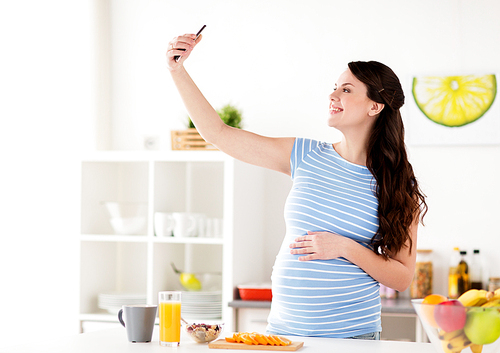 pregnancy, people and technology concept - happy pregnant woman with smartphone taking selfie and having breakfast at home kitchen