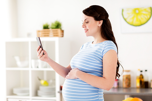 pregnancy, people and technology concept - happy pregnant woman with smartphone taking selfie at home kitchen