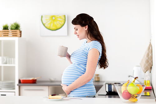 pregnancy, people and expectation concept - happy pregnant woman with cup of tea and piece of cake at home kitchen