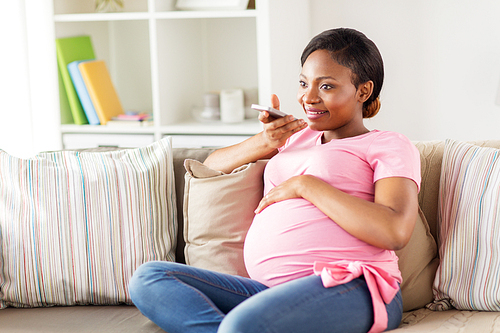pregnancy, technology and people concept - happy pregnant african american woman using voice command recorder or calling on smartphone at home