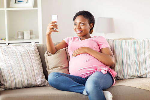 pregnancy, people and technology concept - happy pregnant african american woman with smartphone taking selfie at home