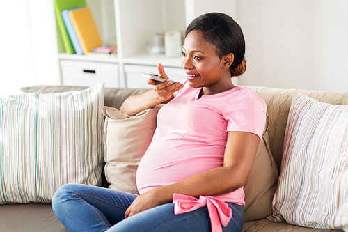 pregnancy, technology and people concept - happy pregnant african american woman using voice command recorder or calling on smartphone at home
