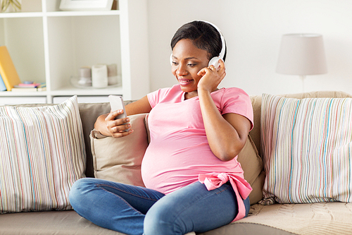 pregnancy, technology and people concept - happy pregnant african american woman with headphones and smartphone listening to music at home