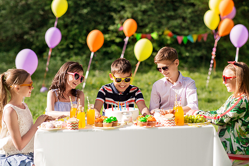 holidays, childhood and celebration concept - happy kids with candles on birthday cake sitting at table at summer garden party