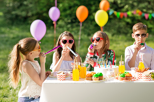 holidays, childhood and celebration concept - happy kids with cake blowing party horns and having fun at summer birthday