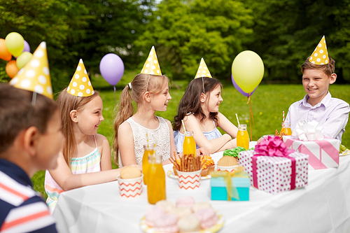 holidays, childhood and celebration concept - happy kids sitting at table on birthday party at summer garden