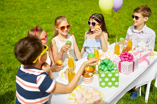 holidays, childhood and celebration concept - happy kids in sunglasses sitting at table on birthday party at summer garden and eating cupcakes