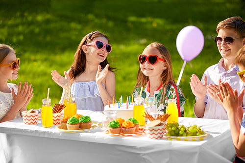 holidays, childhood and celebration concept - happy kids with candles on cake sitting at table at summer garden party and applauding to birthday girl