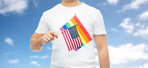 lgbt, same-sex relationships and homosexual concept - close up of man wearing gay pride rainbow awareness wristband and holding american flag over blue sky and clouds background