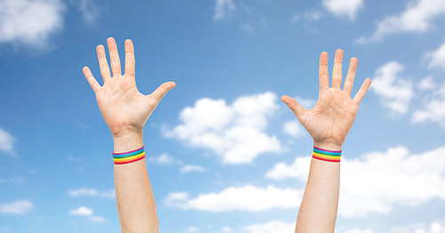 lgbt, same-sex relationships and homosexual concept - close up of male hands wearing gay pride awareness wristbands over blue sky and clouds background