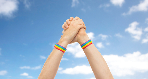 lgbt, same-sex relationships and homosexual concept - close up of male hands wearing gay pride awareness wristbands making winning gesture over blue sky and clouds background