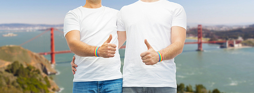 lgbt, same-sex relationships and homosexual concept - close up of male couple with gay pride rainbow awareness wristbands and showing thumbs up over golden gate bridge in san francisco bay background