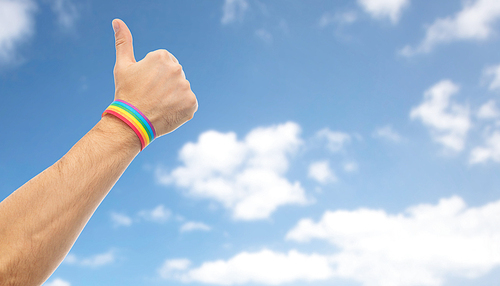 lgbt, same-sex relationships and homosexual concept - close up of male hand wearing gay pride awareness wristband showing thumbs up over blue sky and clouds background