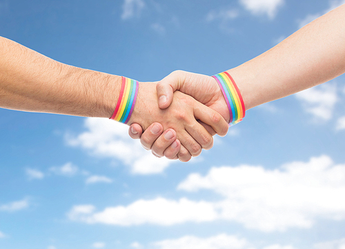 lgbt, same-sex love and homosexual relationships concept - close up of male couple hands with gay pride rainbow awareness wristbands making handshake over blue sky and clouds background