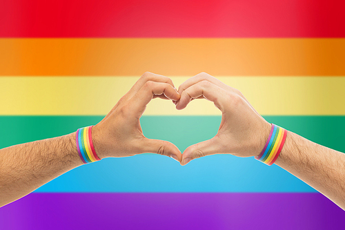 lgbt, same-sex love and homosexual relationships concept - close up of male couple hands with gay pride rainbow awareness wristbands showing heart gesture