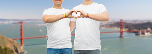 lgbt, same-sex love and homosexual relationships concept - close up of male couple with gay pride rainbow awareness wristbands showing heart gesture over golden gate bridge in san francisco background