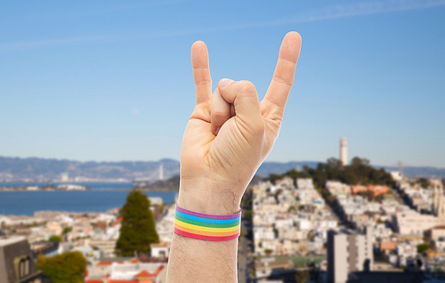 lgbt, same-sex relationships and homosexual concept - close up of male hand wearing gay pride awareness wristband showing rock or hand-horns sign over san francisco city background