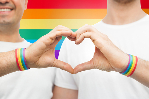 lgbt, same-sex love and homosexual relationships concept - close up of happy smiling male couple wearing gay pride awareness wristbands showing hand heart gesture over rainbow colors background