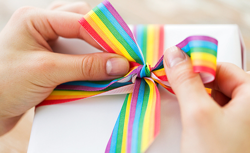 homosexual and lgbt concept - close up of female hands packing gift box and tying bow of gay pride awareness ribbon