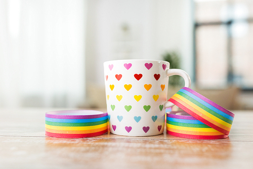 homosexual and lgbt concept - close up of cup with rainbow colored heart pattern and gay pride awareness ribbon on wooden table at home