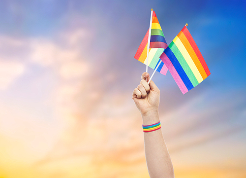 lgbt, same-sex relationships and homosexual concept - close up of male hand with gay pride awareness wristband holding rainbow flags over evening sky background