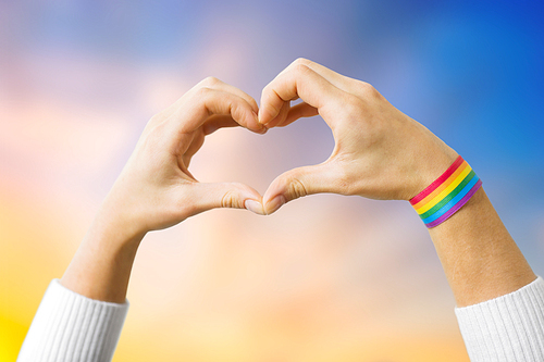 homosexual and lgbt concept - close up of woman wearing gay pride awareness ribbon wristband showing hand heart over evening sky background