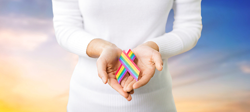 homosexual and lgbt concept - close up of female hands holding gay pride awareness ribbon over evening sky background