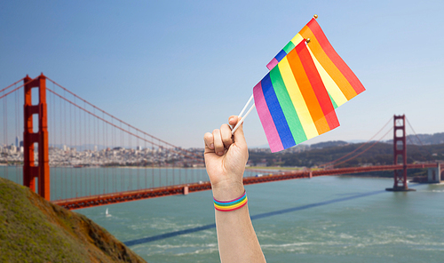 lgbt, same-sex relationships and homosexual concept - close up of male hand with gay pride awareness wristband holding rainbow flags over golden gate bridge in san francisco bay background