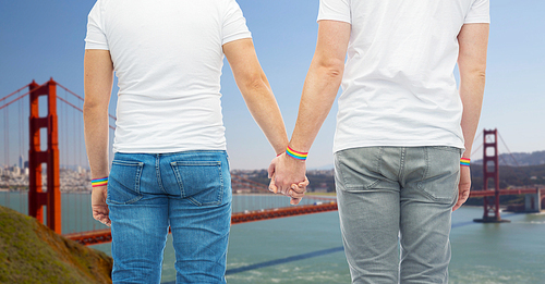 lgbt, same-sex relationships and homosexual concept - close up of male couple wearing gay pride rainbow awareness wristbands holding hands over golden gate bridge in san francisco bay background