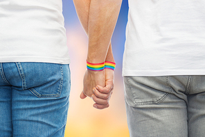 lgbt, same-sex relationships and homosexual concept - close up of male couple wearing gay pride  awareness wristbands holding hands over evening sky background