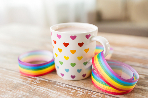homosexual and lgbt concept - close up of cup with rainbow colored heart pattern and gay pride awareness ribbon on wooden table